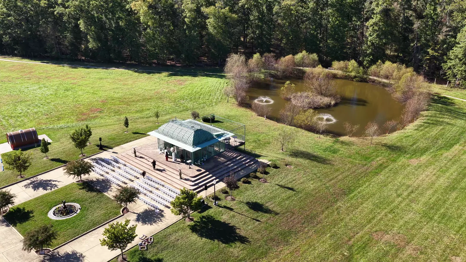 The glass ceremony pavilion from the air, rows of empty white chairs set out in front of it and a fountain pond behind