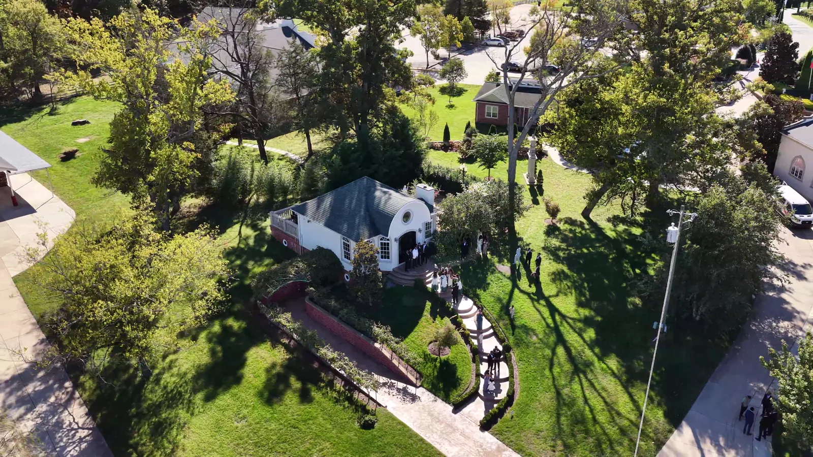 From above: guests on a winding garden path to a small white building among tall trees