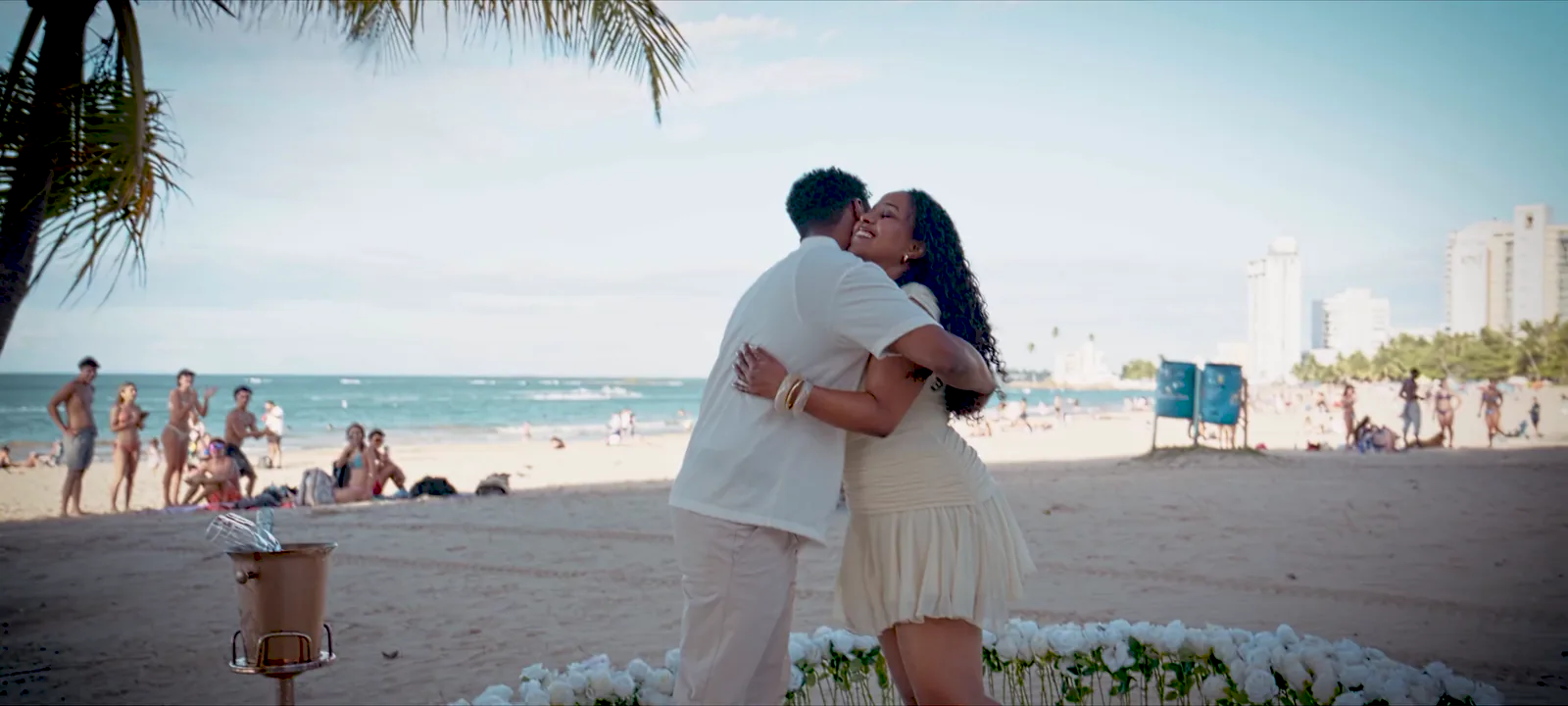 On the beach, the couple embracing inside a ring of white roses, palm trees and the sea behind them