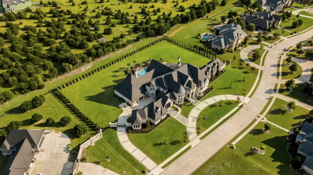 A large stone house on a landscaped lot with a pool and a curved driveway, seen from a drone above the neighborhood.
