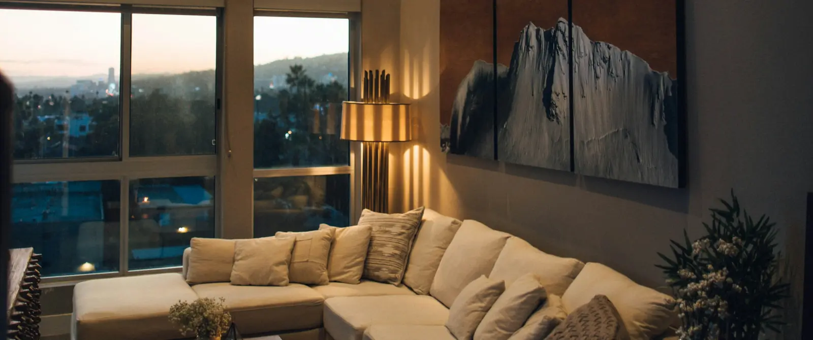 A living room at dusk, framed as a wide film shot: city lights through tall windows, a lit floor lamp, a painting and a sofa.