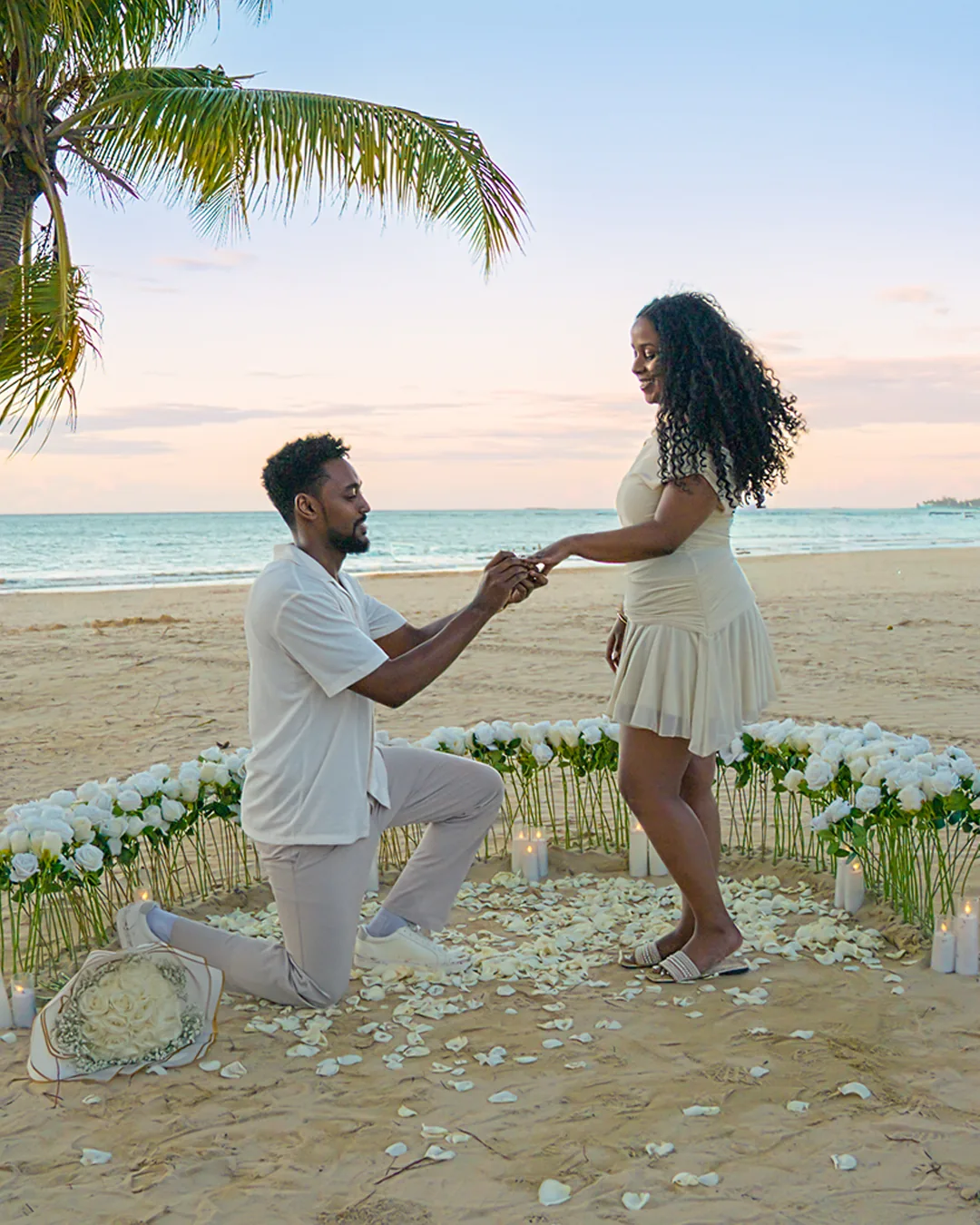 On one knee inside a ring of white roses and candles on the sand, sliding the ring onto a finger