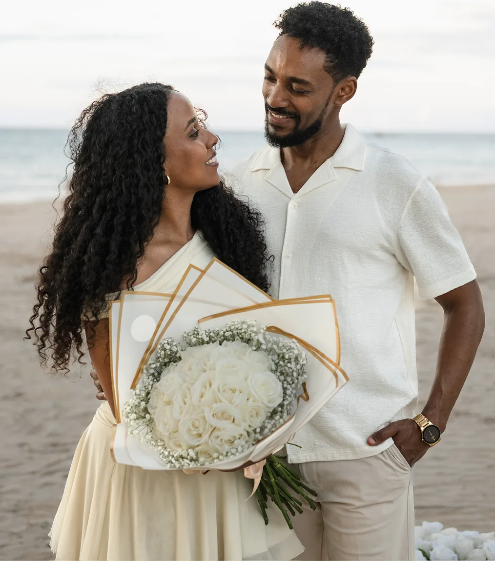 One of the couple holding a wrapped bouquet of white roses, the other close beside, both smiling on the beach