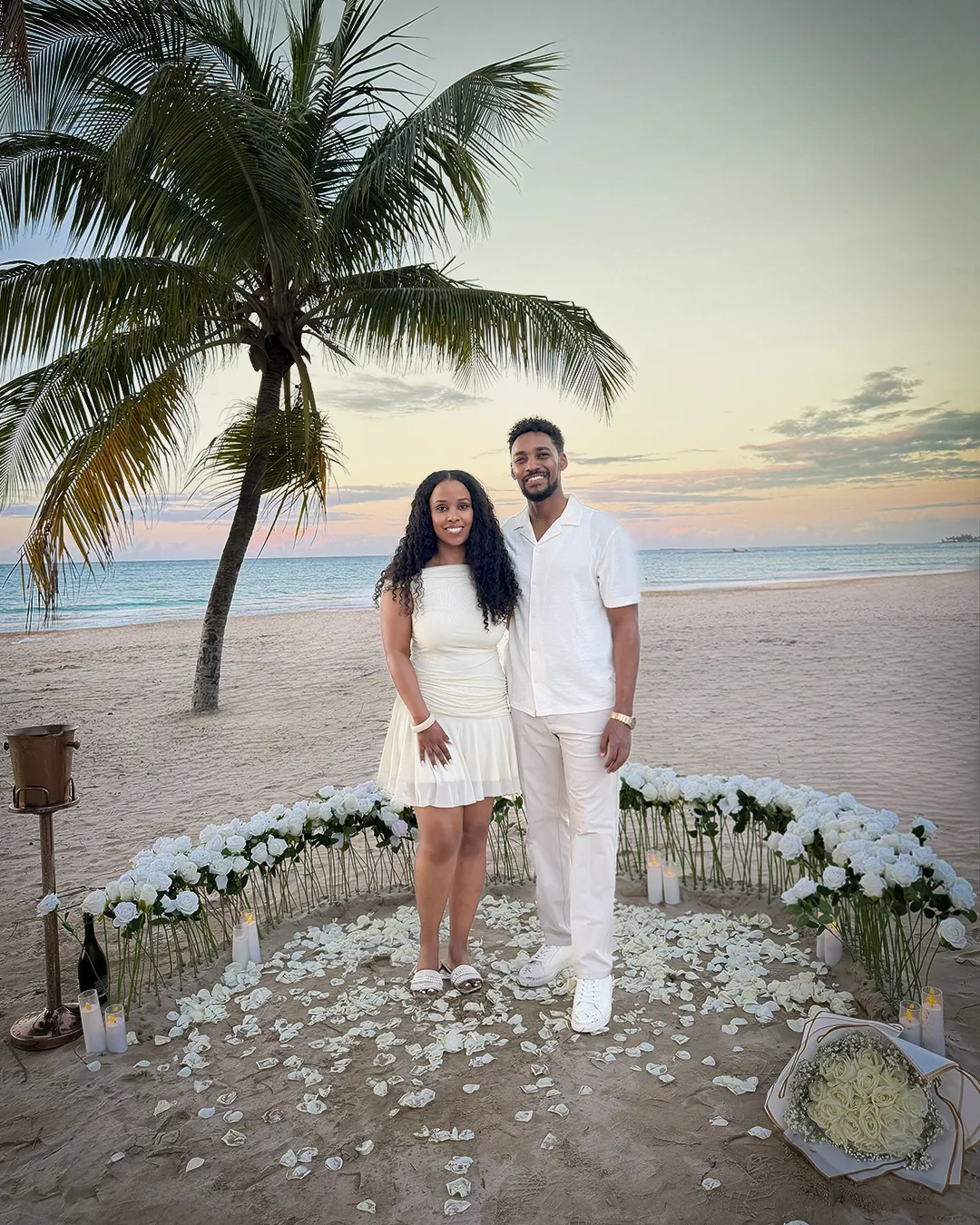 The couple standing together inside the ring of white roses, a palm tree and the sea behind them