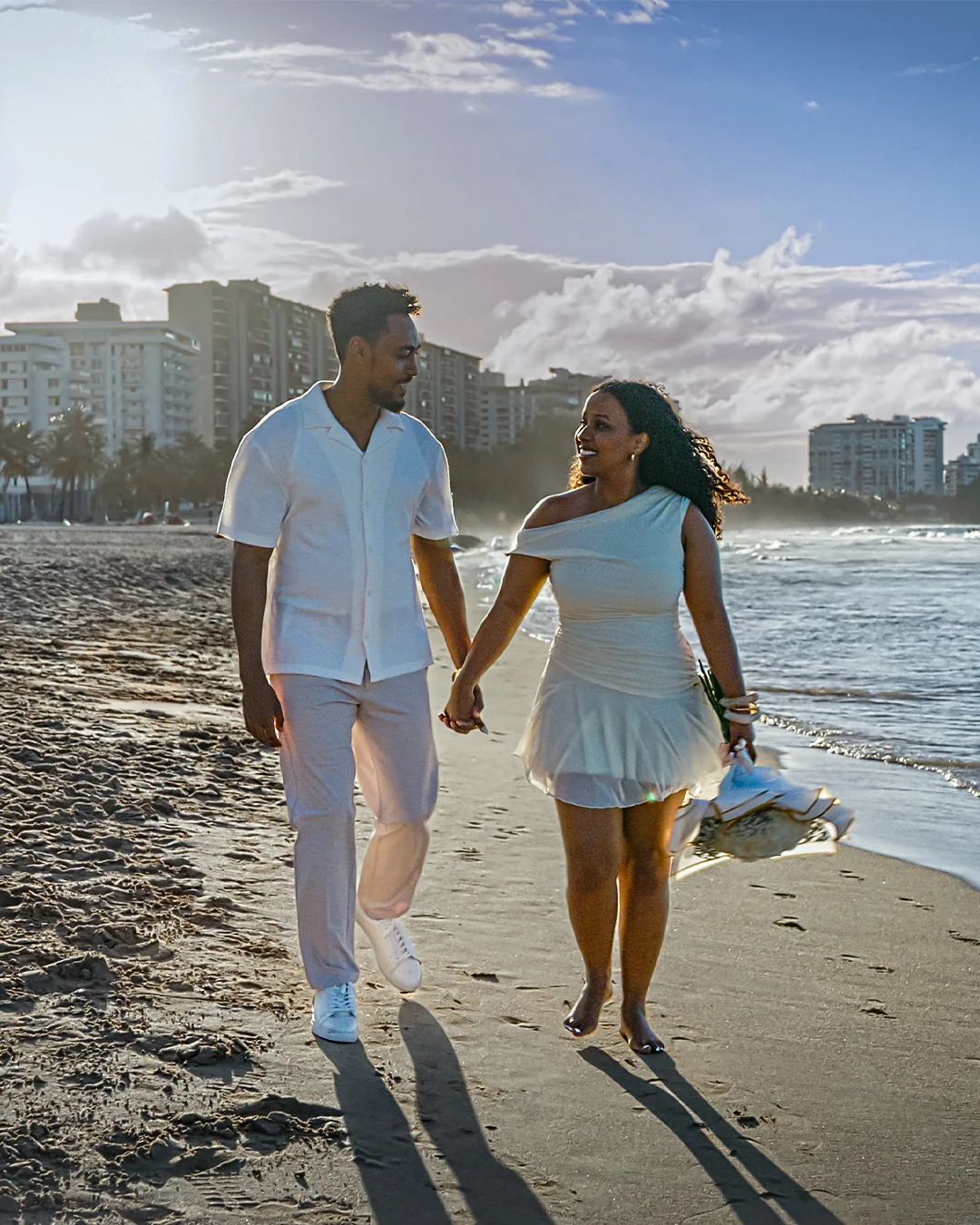 The couple walking hand in hand along the shoreline, high-rise buildings and a bright sky behind them